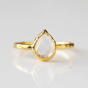 May include: A gold ring with a pear-shaped, iridescent gemstone. The ring's band has a textured, hammered design. The gemstone is set in a gold bezel, and the ring is displayed on a white surface.