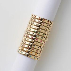 Puede incluir: Una pila de anillos dorados, cada uno adornado con un patrón repetitivo de diseños en forma de estrella. Las estrellas están adornadas con varias piedras preciosas de colores, incluyendo rojo, verde, azul y amarillo, creando una exhibición vibrante y llamativa.