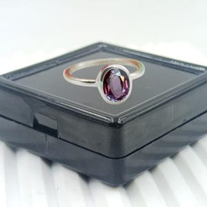 Puede incluir: Un anillo de plata con una piedra preciosa ovalada de color púrpura oscuro. El anillo está colocado sobre una caja de joyería negra y cuadrada, que a su vez está sobre una superficie blanca y estriada. La piedra preciosa tiene una superficie pulida y reflectante.