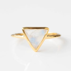 Pode incluir: Um anel dourado com uma pedra preciosa triangular e iridescente. A pedra tem uma base branca com tons azuis sutis. A banda e a cravação do anel são douradas, e a banda tem um design texturizado.