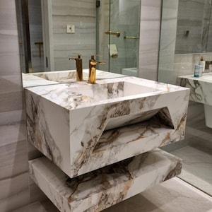 Peut inclure: Un lavabo mural moderne avec un motif marbré blanc, marron et doré. Le lavabo a un bassin rectangulaire et des robinets dorés. La conception comprend une étagère inférieure. La pièce comprend un grand miroir.