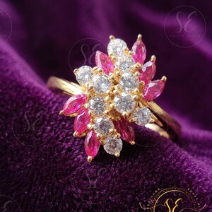Puede incluir: Un anillo de oro con un grupo de piedras redondas, transparentes y facetadas en el centro, rodeadas de piedras de color rosa vibrante con talla marquesa. Las piedras están engastadas en una banda de metal dorado que sostiene el diseño floral.