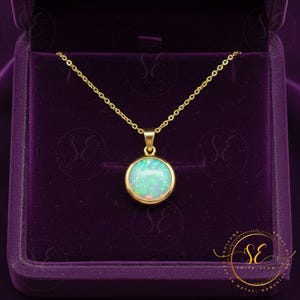 Könnte beinhalten: Eine goldfarbene Halskette mit einem runden Anhänger, der einen hellblauen und weißen Opalstein zeigt. Der Anhänger ist in einer goldenen Lünette gefasst und hängt an einer zarten Goldkette. Die Halskette wird in einer violetten Samt-Schmuckschatulle präsentiert.