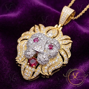 May include: A gold lion pendant necklace with intricate detailing. The lion's head is adorned with small, clear stones and has red gemstone eyes and a ruby at the base. The pendant is suspended from a fine gold chain.