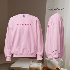 Könnte beinhalten: Hellrosa Sweatshirt mit dem Wort "nurse" in Schreibschrift auf der Brust gestickt. Das Wort "Embroidered" ist in Schreibschrift oben rechts geschrieben. Auf dem Ärmel befindet sich ein kleines Herzdesign.