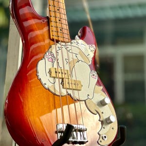 Może przedstawiać: Gitara basowa z wykończeniem sunburst, czerwonym, pomarańczowym i brązowym korpusem. Gitara ma wzór kota z kreskówek. Szyjka jest jasnobrązowa, struny są widoczne. Stoi na czarnym stojaku.