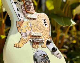 May include: A mint green electric guitar with a jaguar graphic. The guitar has a chrome bridge and black knobs. The fretboard is light brown. The guitar is displayed on a black stand, with green foliage in the background.