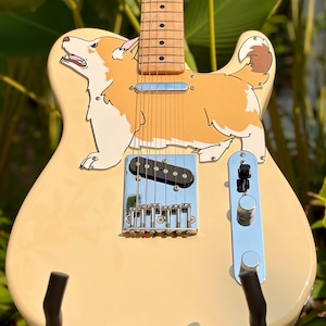 Puede incluir: Una guitarra eléctrica de color crema con una gran ilustración gráfica de un perro Corgi de color canela y blanco en el cuerpo. El instrumento incluye un puente cromado, una pastilla de bobina simple, dos perillas de control plateadas y un mástil de arce.