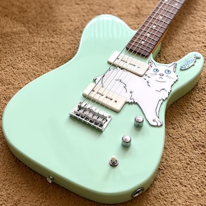 May include: A mint green electric guitar featuring a custom white pickguard shaped like a fluffy white cat with blue eyes. The instrument includes two cream-coloured soapbar pickups, a chrome bridge, three silver control knobs, and a small fish sticker.