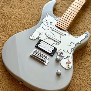 May include: A light gray electric guitar with a white cat graphic. The guitar features a maple neck, black and white pickups, and silver hardware. The cat graphic has pink paw pads.