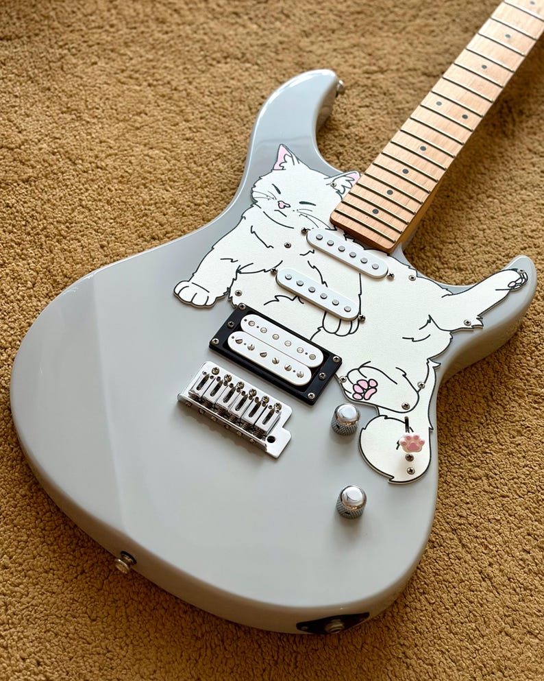 May include: A light gray electric guitar with a white cat graphic. The guitar features a maple neck, black and white pickups, and silver hardware. The cat graphic has pink paw pads.