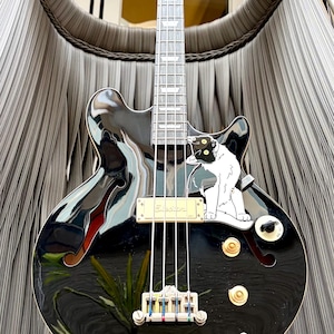 May include: A black electric bass guitar with a cat decal. The guitar features a hollow body design, f-holes, gold knobs, and a silver bridge. The fretboard is silver and the strings are white. The guitar's body reflects the surrounding environment.