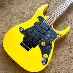Op de afbeelding: Felgele elektrische gitaar met een zwarte kattenafbeelding. De gitaar heeft zwarte hardware, inclusief de brug en pickups. De toets is lichtgekleurd en heeft twee gele volume- en toonregelaars. Modern design.