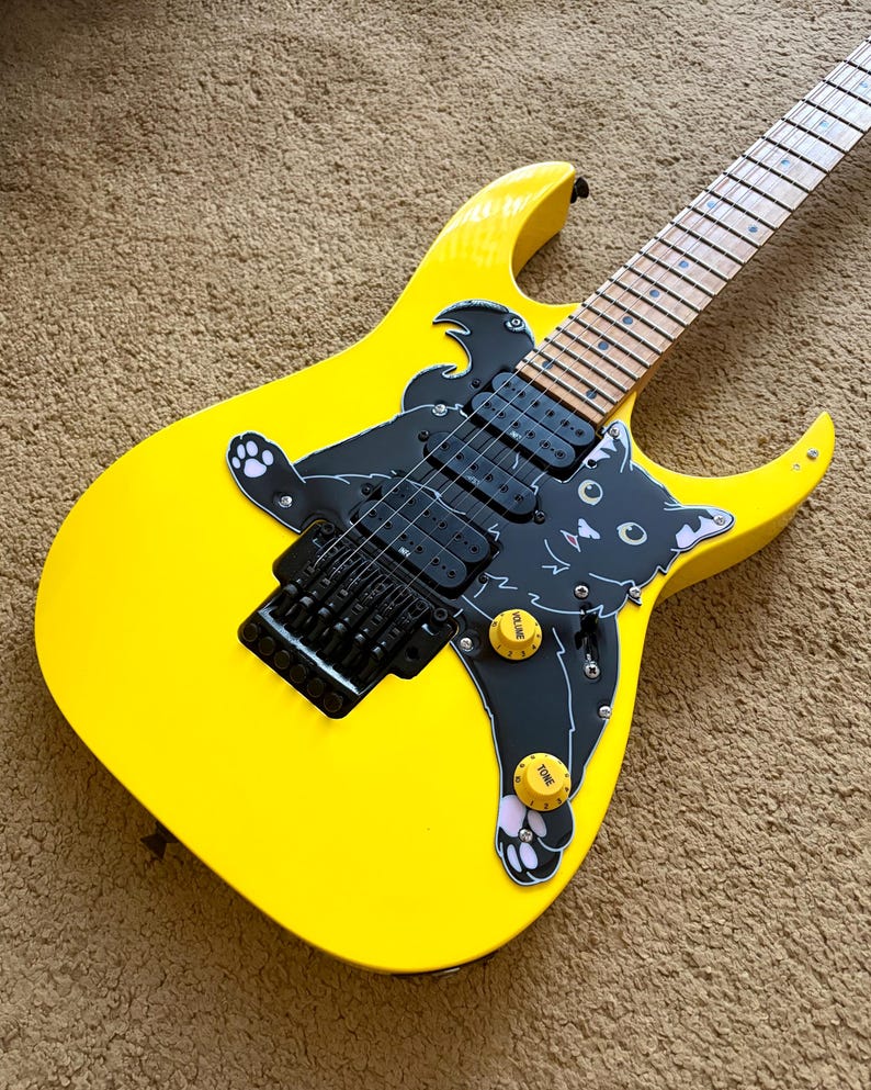 Op de afbeelding: Felgele elektrische gitaar met een zwarte kattenafbeelding. De gitaar heeft zwarte hardware, inclusief de brug en pickups. De toets is lichtgekleurd en heeft twee gele volume- en toonregelaars. Modern design.