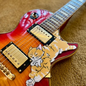Puede incluir: Una guitarra eléctrica con acabado sunburst y un diseño de gato de dibujos animados. La guitarra presenta pastillas y herrajes dorados. El gráfico del gato es naranja y blanco, y está durmiendo en el cuerpo de la guitarra.