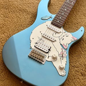 May include: A light blue electric guitar featuring a custom white pickguard with a graphic illustration of a white cat and a small fish. The instrument includes a dark fretboard, silver bridge hardware, and two white volume and tone control knobs.