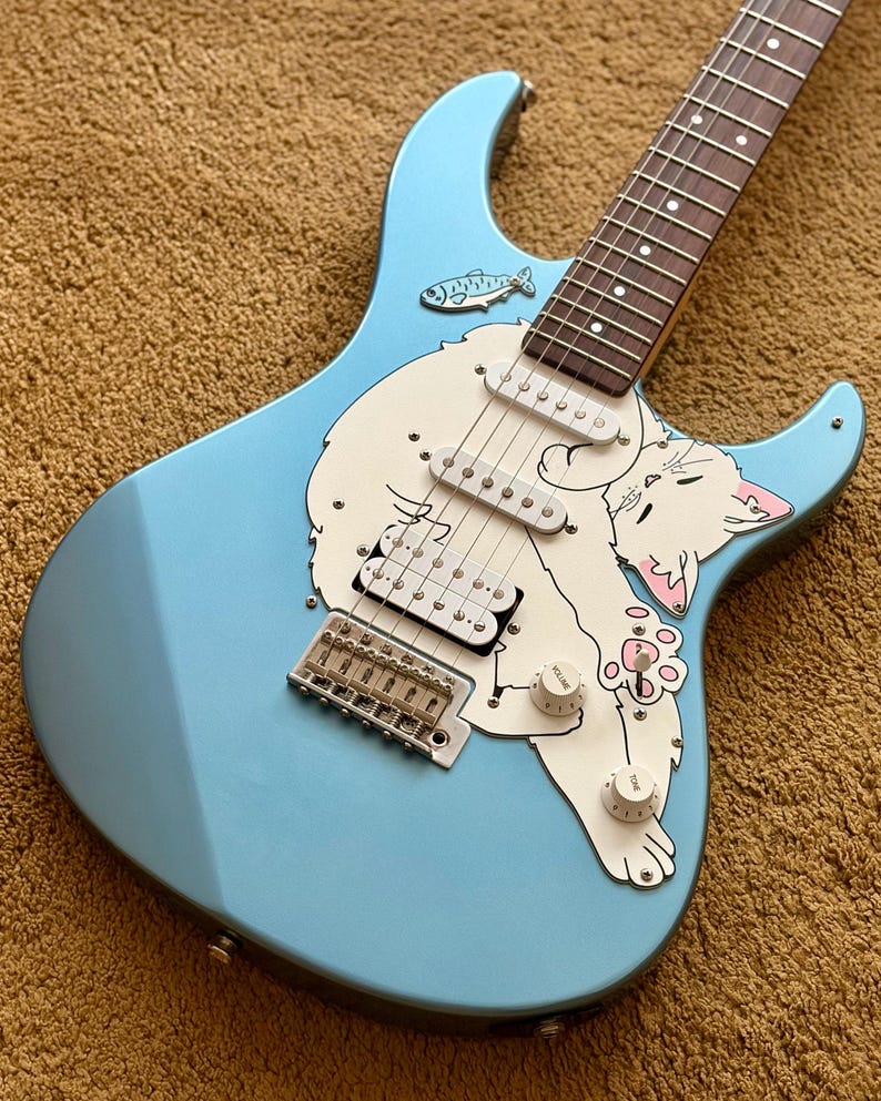 May include: A light blue electric guitar featuring a custom white pickguard with a graphic illustration of a white cat and a small fish. The instrument includes a dark fretboard, silver bridge hardware, and two white volume and tone control knobs.