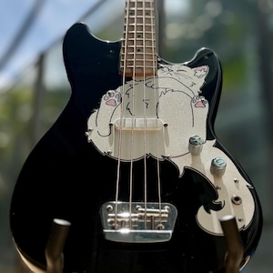 May include: A black electric bass guitar with a white cat graphic on the body. The cat illustration features pink paw pads. The guitar has silver strings, knobs, and a bridge. The fretboard is light brown.