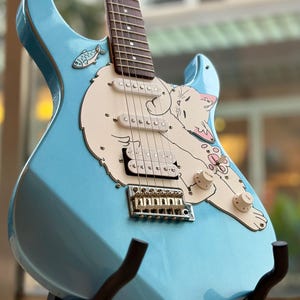 May include: A light blue electric guitar featuring a cream-colored pickguard shaped like a sleeping cat. The guitar includes three pickups, a chrome bridge, and a small fish graphic near the neck. The body has a glossy finish with volume and tone knobs.