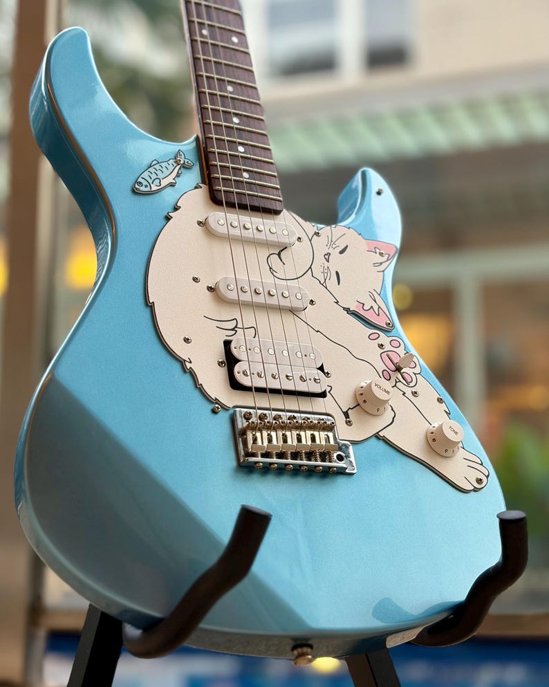 May include: A light blue electric guitar featuring a cream-colored pickguard shaped like a sleeping cat. The guitar includes three pickups, a chrome bridge, and a small fish graphic near the neck. The body has a glossy finish with volume and tone knobs.