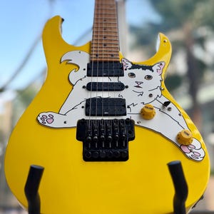 Op de afbeelding: Een gele elektrische gitaar met een witte kattenafbeelding. De gitaar heeft zwarte pickups, een tremolo brug en gele volume- en toonregelaars. De kat heeft roze voetzolen. De gitaar staat op een zwarte standaard.