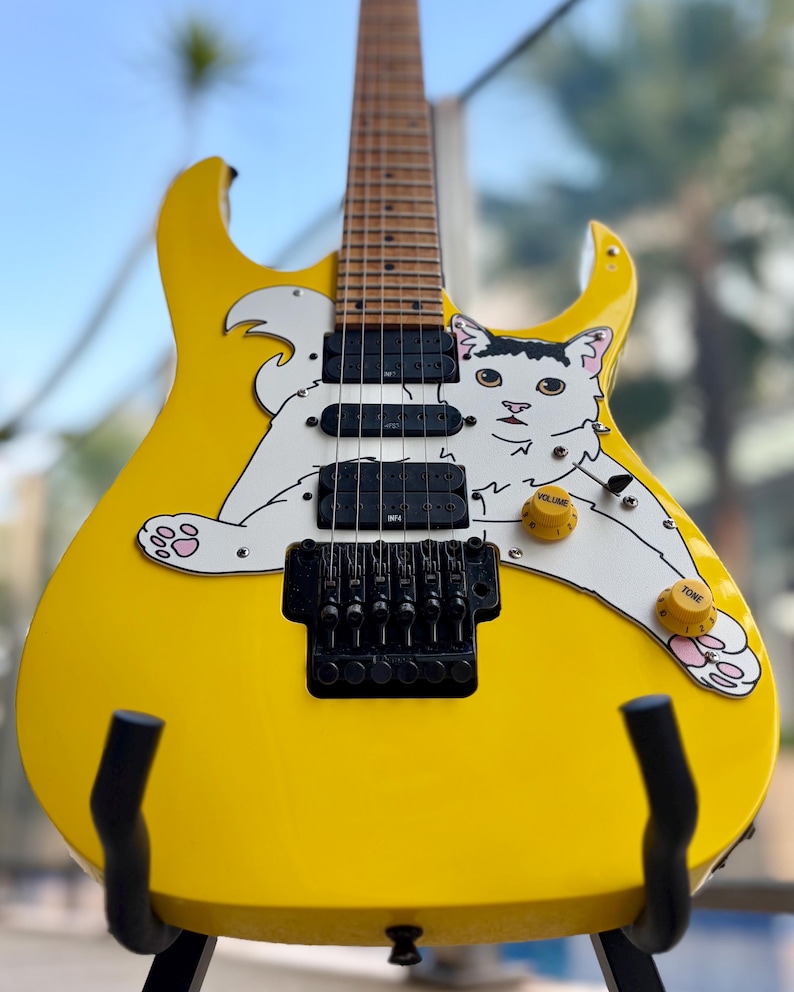Op de afbeelding: Een gele elektrische gitaar met een witte kattenafbeelding. De gitaar heeft zwarte pickups, een tremolo brug en gele volume- en toonregelaars. De kat heeft roze voetzolen. De gitaar staat op een zwarte standaard.