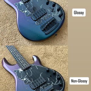 Op de afbeelding: Een basgitaar met een glanzende en niet-glanzende afwerking. De body heeft een verloop van paars naar teal. Een zwarte slagplaat heeft een witte illustratie. De gitaar heeft zwarte hardware en snaren.