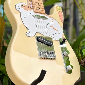 May include: A cream-coloured electric guitar featuring a large white and brown rabbit graphic on the body, a single orange carrot illustration, a maple neck, chrome hardware, a bridge pickup, and two silver control knobs on a metal plate.