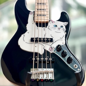 Könnte beinhalten: Ein schwarzer elektrischer Bass mit einem individuellen weißen Schlagbrett in Form einer flauschigen weißen Katze mit blauen Augen. Das Instrument hat einen hellen Holzhals, schwarze Tonabnehmer, drei schwarze Regler und eine Chrombrücke.