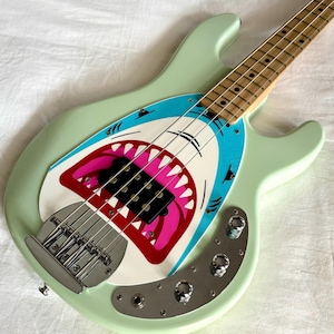 Puede incluir: Un bajo eléctrico verde menta con un diseño de boca de tiburón en el golpeador. La boca del tiburón es blanca, azul y rosa, con afilados dientes blancos. El bajo tiene un mástil de madera y herrajes plateados.