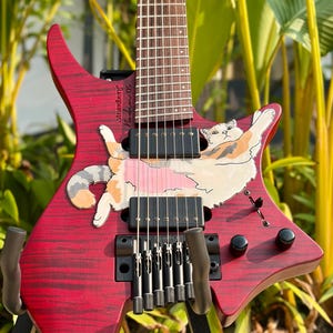 May include: A headless Strandberg Boden OS electric guitar with a deep red flamed maple finish. The body features a large, hand-painted illustration of a calico cat stretched across the center. It includes black hardware, two humbucker pickups, and a dark fretboard.
