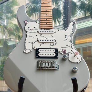 May include: A gray electric guitar featuring a playful white cat design. The guitar has a wooden neck, white pickups, and chrome hardware. The cat illustration includes pink accents on the ears and paws, adding a whimsical touch to the instrument.