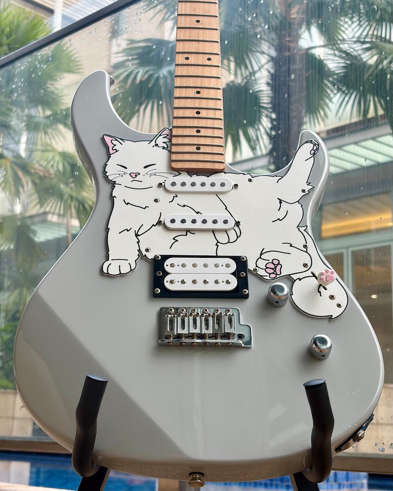 May include: A gray electric guitar featuring a playful white cat design. The guitar has a wooden neck, white pickups, and chrome hardware. The cat illustration includes pink accents on the ears and paws, adding a whimsical touch to the instrument.