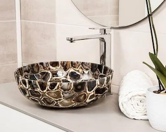 May include: A decorative, round bathroom sink made of brown, gray, and white agate stones. The sink sits on a light gray countertop next to a chrome faucet, a rolled white towel, and a potted plant.