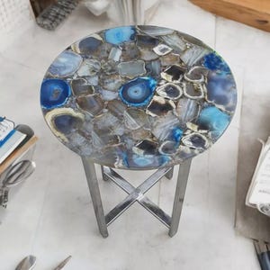 May include: A round side table with a mosaic top made of blue, gray, and white agate stones. The table has a silver metal base with crossed legs. The table is approximately 18 inches in diameter.