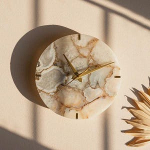 May include: A round wall clock with a natural stone face, featuring shades of beige, cream, and brown. The clock has gold-colored hands and minimalist black markers. The clock is mounted on a light beige wall.