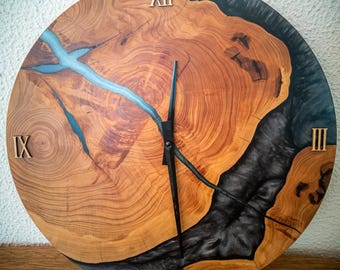May include: A round wooden clock with a natural wood grain pattern. The clock face features Roman numerals at the 3, 6, 9, and 12 o'clock positions. A dark blue resin fills the gaps in the wood, and the clock hands are black.
