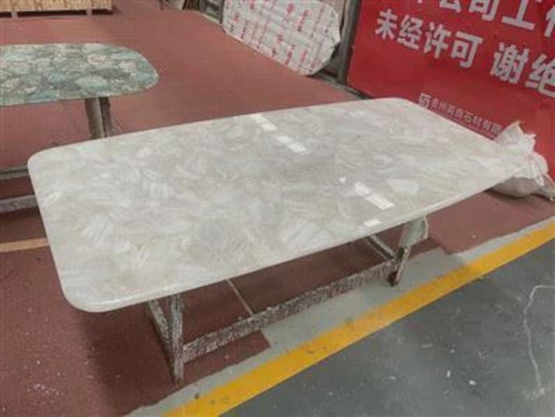 Pu&ograve; includere: Un tavolo rettangolare con una superficie chiara e decorata. Il tavolo ha una base grigio chiaro ed &egrave; posto su una superficie rossa con una striscia gialla. Un altro tavolo con una superficie verde &egrave; visibile sullo sfondo.
