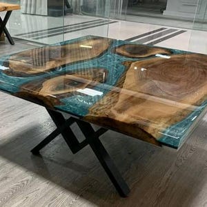 Peut inclure: Table basse carrée avec un dessus en résine époxy transparente et des inclusions de grain de bois naturel. La résine est de couleur sarcelle et la table a un piètement en métal noir. La table mesure environ 122 cm de long et de large.