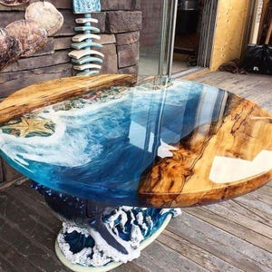 Pode incluir: Uma mesa redonda artesanal com um design temático do oceano. A mesa apresenta um tampo de resina transparente com um padrão de ondas azuis e brancas, uma estrela do mar e uma borda de madeira. A base é um design de onda esculpida.