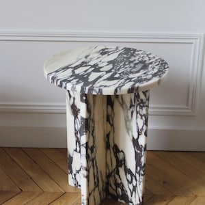 May include: A round marble side table with a white and black veined pattern. The table has a cylindrical base with a cross-shaped support. The table is approximately 50 cm in diameter and 50 cm tall.