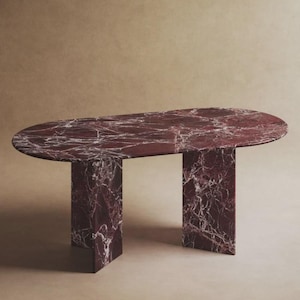 Op de afbeelding: Een moderne ovale tafel met een bordeauxrode marmeren blad en twee rechthoekige poten. Het marmer heeft witte aders. Een design meubelstuk voor eetkamer of woonkamer.