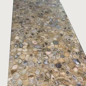 Puede incluir: Una superficie rectangular cubierta por un mosaico de piedras pulidas de forma irregular. Las piedras son principalmente beige, grises y marrones, algunas con patrones concéntricos. La superficie tiene un acabado brillante y una estética natural y orgánica.