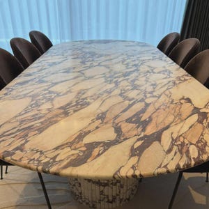 May include: An oval marble dining table with a cream and brown patterned surface. The table is surrounded by dark brown upholstered chairs. The table's base is a cylindrical design made of marble. The table is approximately 254 cm long.