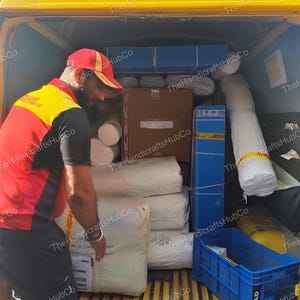 May include: A delivery worker in a red and yellow uniform loads white fabric rolls and cardboard boxes into the back of a yellow van. A blue plastic crate sits on the floor of the vehicle, which is filled with various packages and shipping materials.