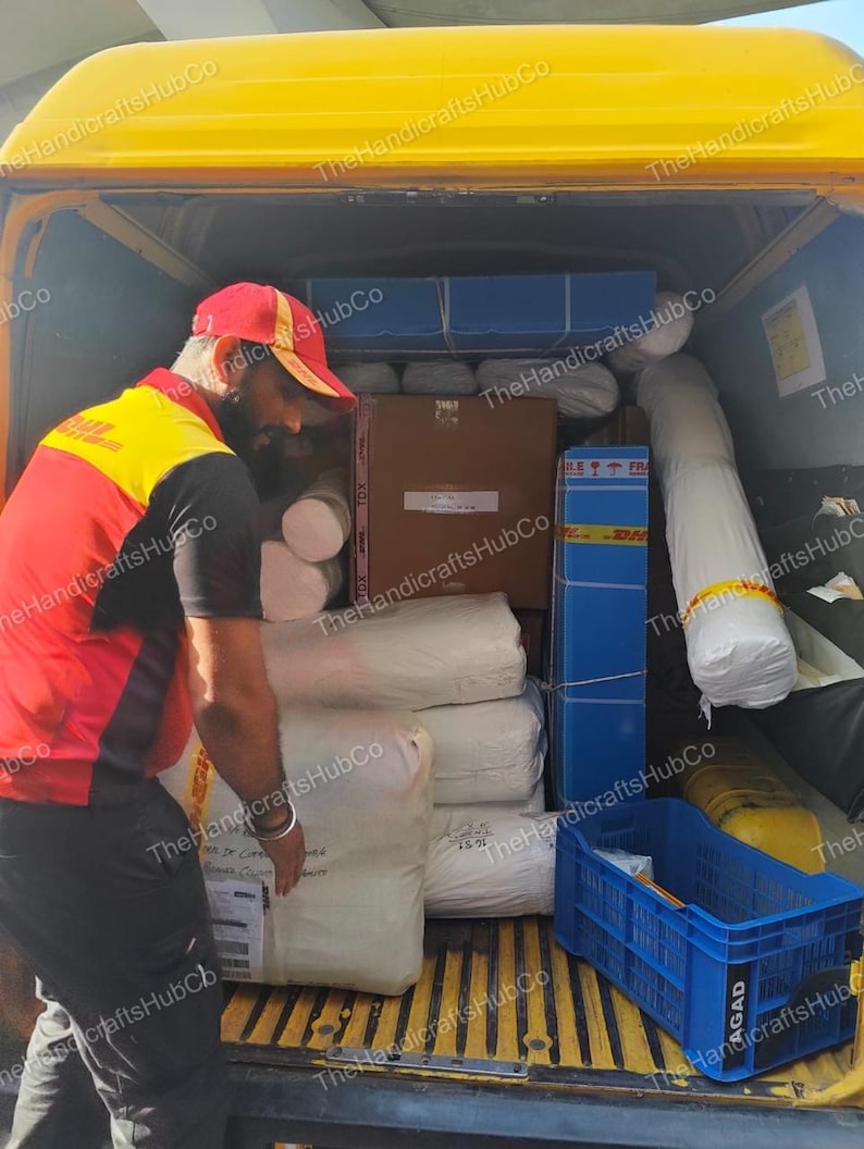 May include: A delivery worker in a red and yellow uniform loads white fabric rolls and cardboard boxes into the back of a yellow van. A blue plastic crate sits on the floor of the vehicle, which is filled with various packages and shipping materials.