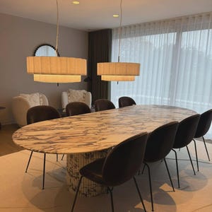 Puede incluir: Un comedor moderno con una gran mesa ovalada de mármol y sillas de terciopelo oscuro. Dos lámparas colgantes en niveles cuelgan sobre la mesa. La habitación tiene una paleta de colores neutros, con una alfombra clara.