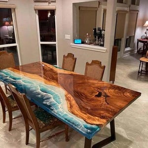 Peut inclure: Une table de salle à manger rectangulaire au design unique. La table présente une surface en bois brillant avec un motif en résine bleue et blanche ressemblant à des vagues océaniques. Elle est soutenue par un piètement en métal foncé et entourée de chaises en bois.