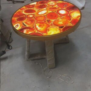 May include: A round side table featuring a backlit tabletop composed of numerous polished orange and red agate slices. The table has a thin gold-toned metal rim and a base wrapped in a textured, light-colored fibrous material with a power cord.