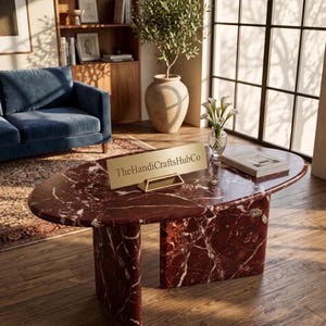 May include: An oval coffee table made of deep red marble with prominent white veining, featuring two thick, curved slab legs. A gold-toned rectangular sign with the text TheHandiCraftsHubCo is placed on the surface next to a book and a glass vase.